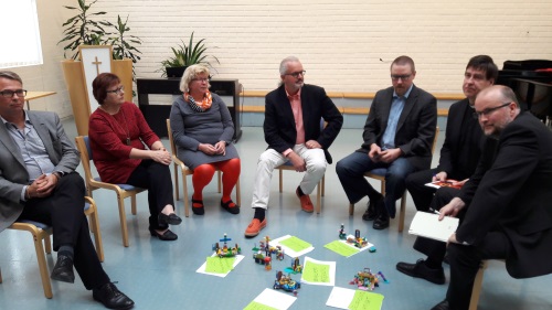 Seitsemän henkilöä istuu puolikaaressa tuoleilla. Lattialla keskellä on papereita ja legorakennelmia.