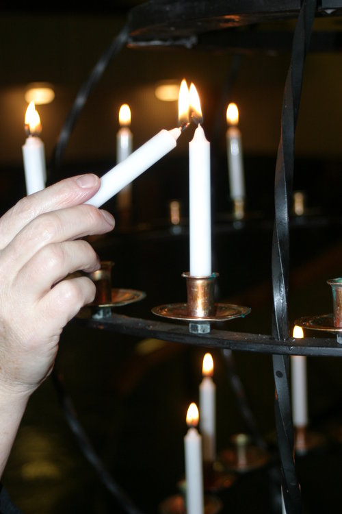 Lähikuva kädestä, joka sytyttää kynttilää kirkon lähetyskynttelikköön. Taustalla muita palavia kynttilöitä.