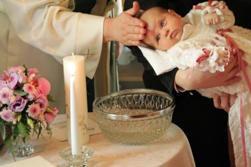 Pappi siunaa juuri kastettua lasta pitämällä kättä lapsen pään päällä. Lapsi on kummin sylissä ja hereillä.
