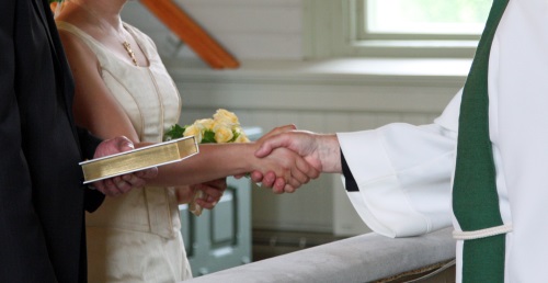 Pappi ojentaa vihitylle parille Raamatun kirkon alttarilla. Lähikuva käsistä ja Raamatusta.